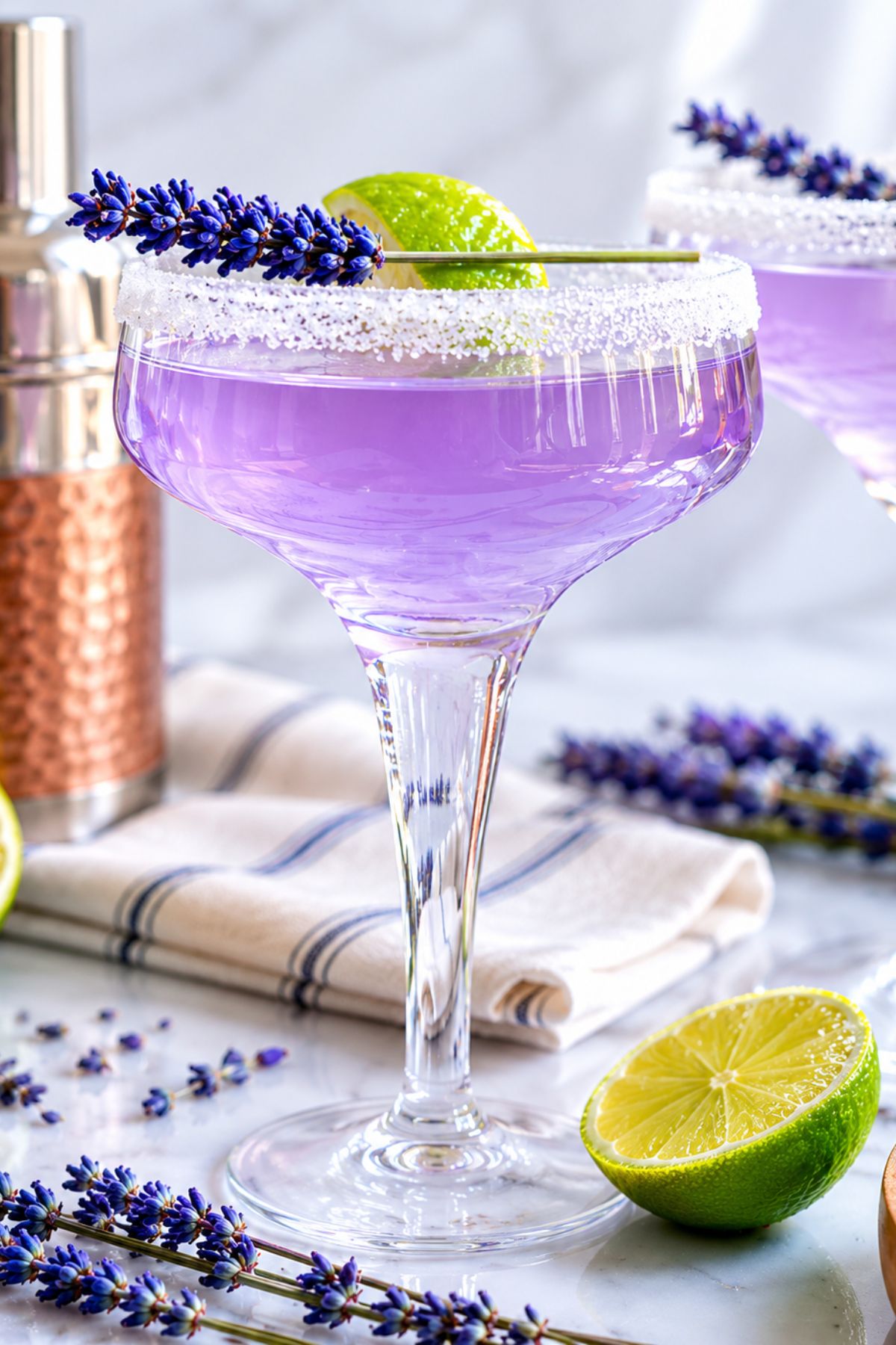 A purple cocktail in a sugared-rim glass, garnished with a lime wheel and lavender sprig, sits on a table with a halved lime, lavender, and a cocktail shaker nearby.
