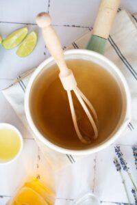 A top-down view of a saucepan filled with light brown liquid, a wooden whisk inside, lime wedges, and a small bowl of juice nearby on a striped cloth.