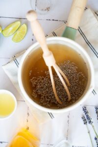 A pot of liquid with dried lavender and a wooden whisk, surrounded by lime wedges, a cup of juice, and a beige cloth on a white surface.