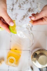A hand rubs a lime wedge on the rim of a margarita glass, with a bottle of liquor and a cocktail shaker nearby on a white surface.