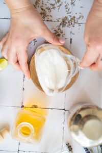 A person rims a glass with sugar on a wooden plate, surrounded by a bottle of yellow liquid, a cocktail shaker, lime wedge, and scattered dried herbs.
