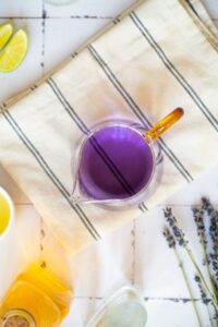 A glass pitcher filled with purple liquid sits on a striped cloth, surrounded by lime wedges, lavender sprigs, and other ingredients on a white surface.