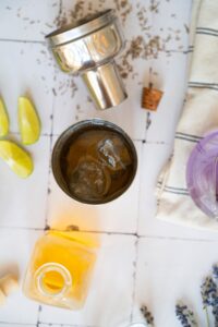 A cocktail shaker with ice is on a tiled surface, surrounded by lime wedges, a metal cap, a striped cloth, a glass bottle, and scattered herbs.