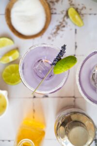 Top view of a purple cocktail with ice, garnished with a lime wedge and lavender sprig, surrounded by lime slices, a bowl of sugar, and ingredients on a tiled surface.