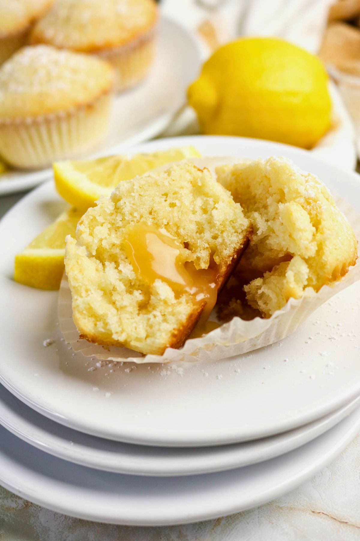 A lemon muffin broken in half on a plate, revealing a gooey lemon filling inside, with lemon slices and whole muffins in the background.