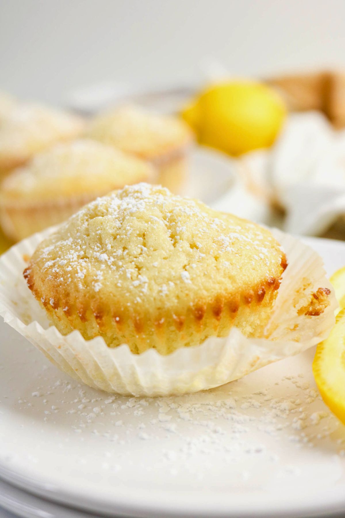 A lemon muffin in a paper liner sits on a white plate, dusted with powdered sugar. Lemon wedges and more muffins are blurred in the background.