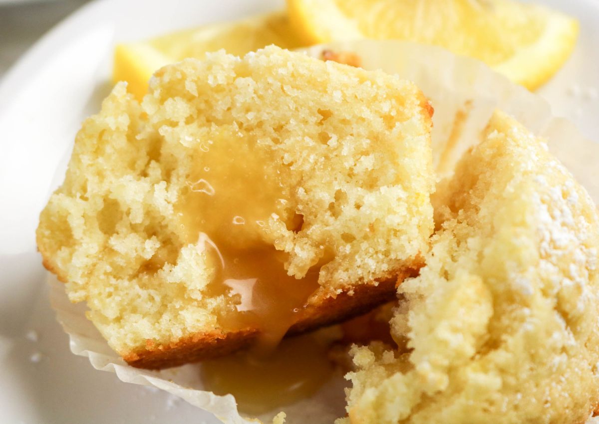 A lemon muffin cut in half on a plate, showing a gooey lemon filling inside, with lemon slices in the background.