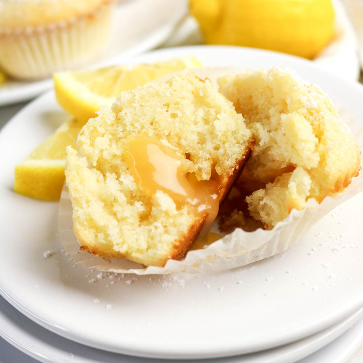 A lemon muffin split open on a white plate, revealing a gooey lemon filling, with lemon slices in the background.