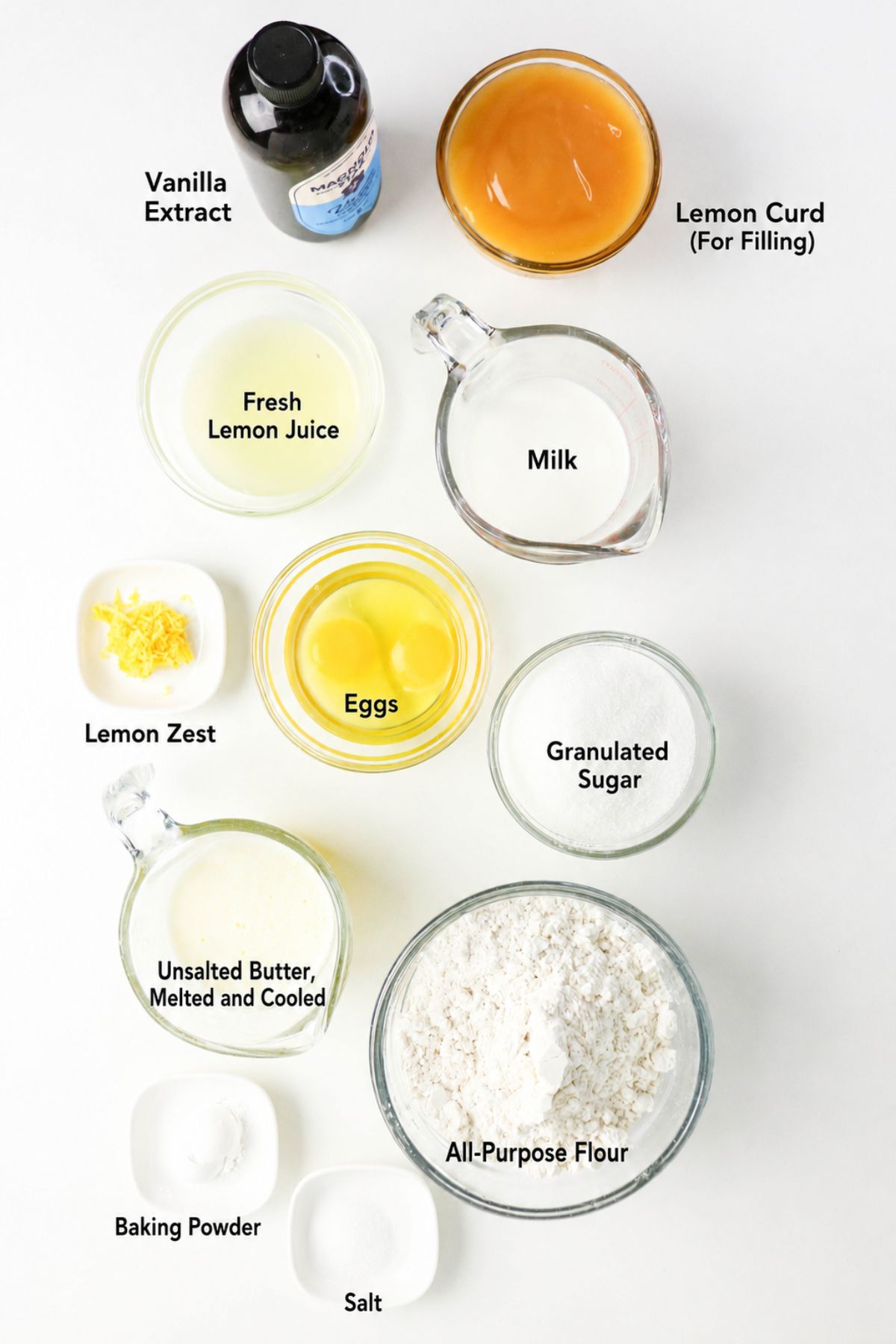 Top-down view of labeled baking ingredients including flour, sugar, eggs, milk, lemon juice, lemon zest, vanilla extract, lemon curd, melted butter, baking powder, and salt on a white surface.