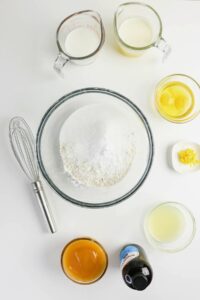 Baking ingredients including flour, sugar, milk, melted butter, lemon juice, lemon zest, vanilla extract, and a whisk arranged on a white surface.