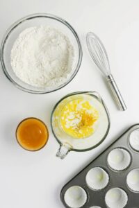 Bowls of flour, melted butter with zest, a cup of orange liquid, a whisk, and a muffin tray with paper liners are arranged on a white surface.