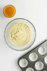 A glass bowl of muffin batter, a glass cup of orange liquid, and a muffin tin lined with paper cups are arranged on a white surface.