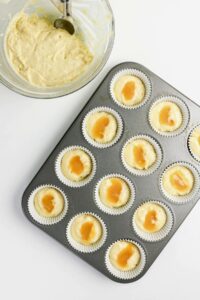A muffin tin with paper liners is filled with batter and topped with dollops of a light brown sauce; a mixing bowl with remaining batter and a spoon is nearby.