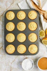 A muffin tin with twelve baked muffins sits on a marble surface beside halved lemons, a bowl of powdered sugar, and a bowl of glaze.