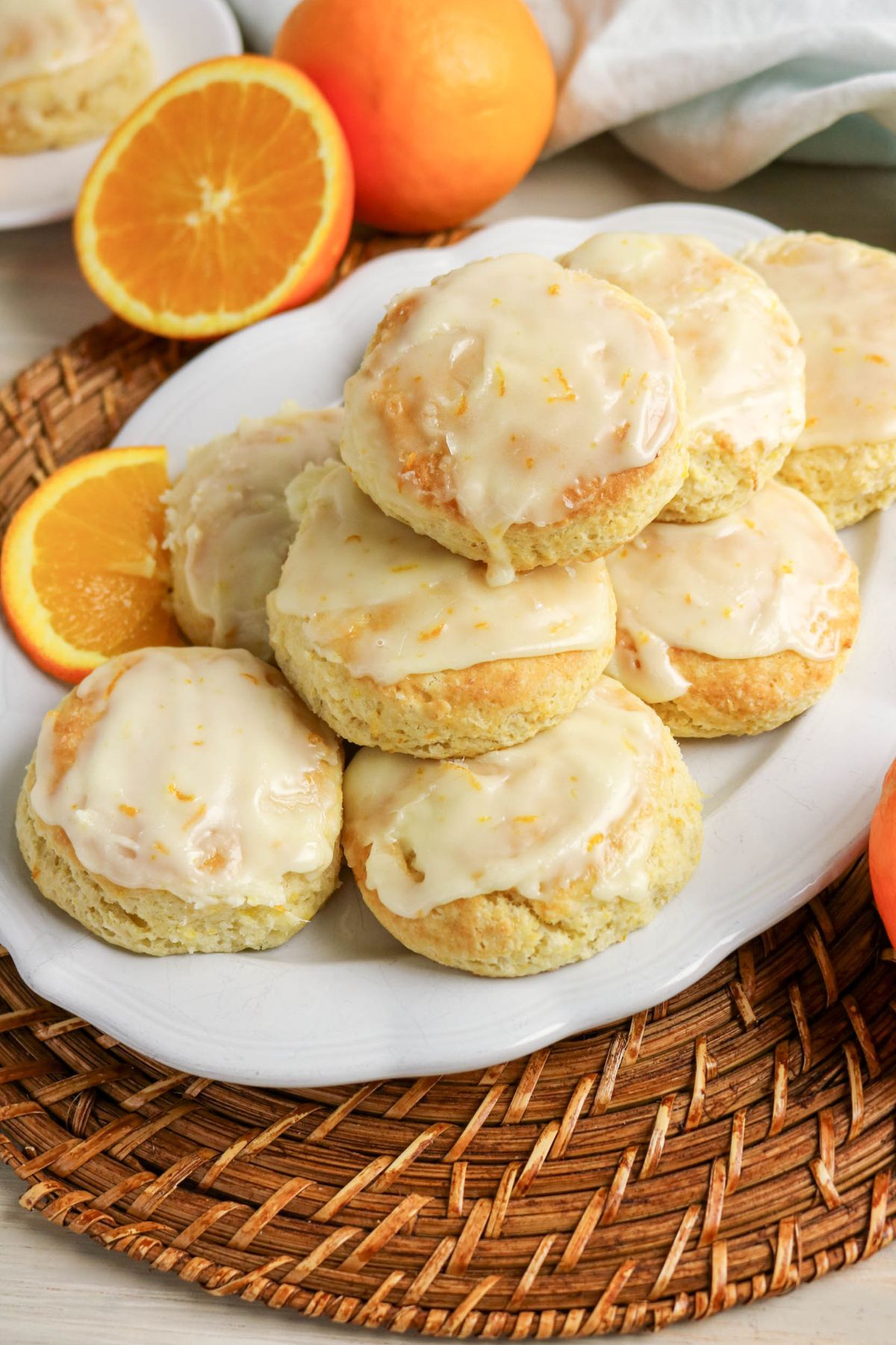A plate of glazed orange scones sits on a woven placemat, accompanied by fresh orange halves and whole oranges in the background.