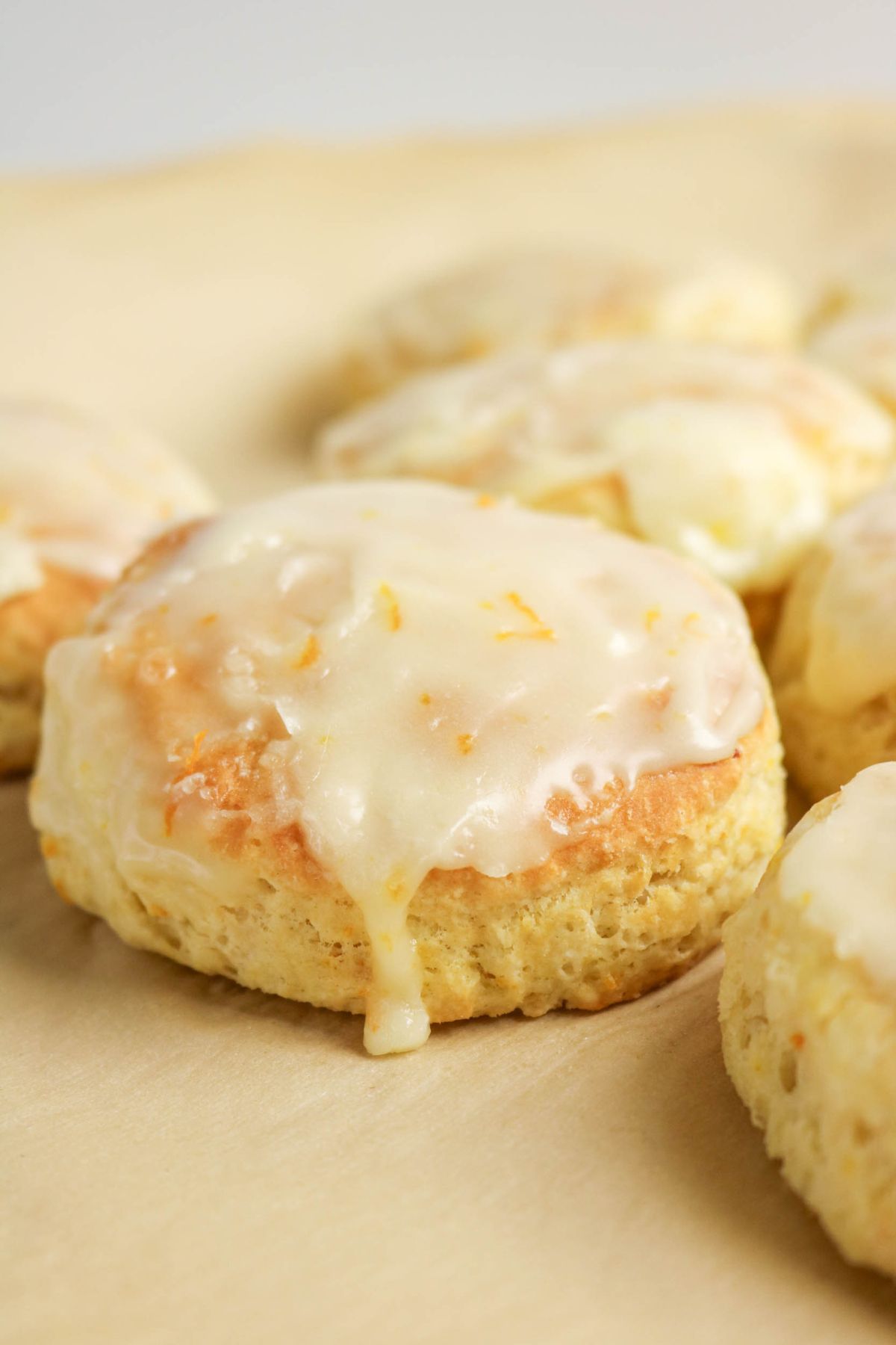 A close-up of a glazed biscuit with icing on top, resting on parchment paper with other similar biscuits in the background.