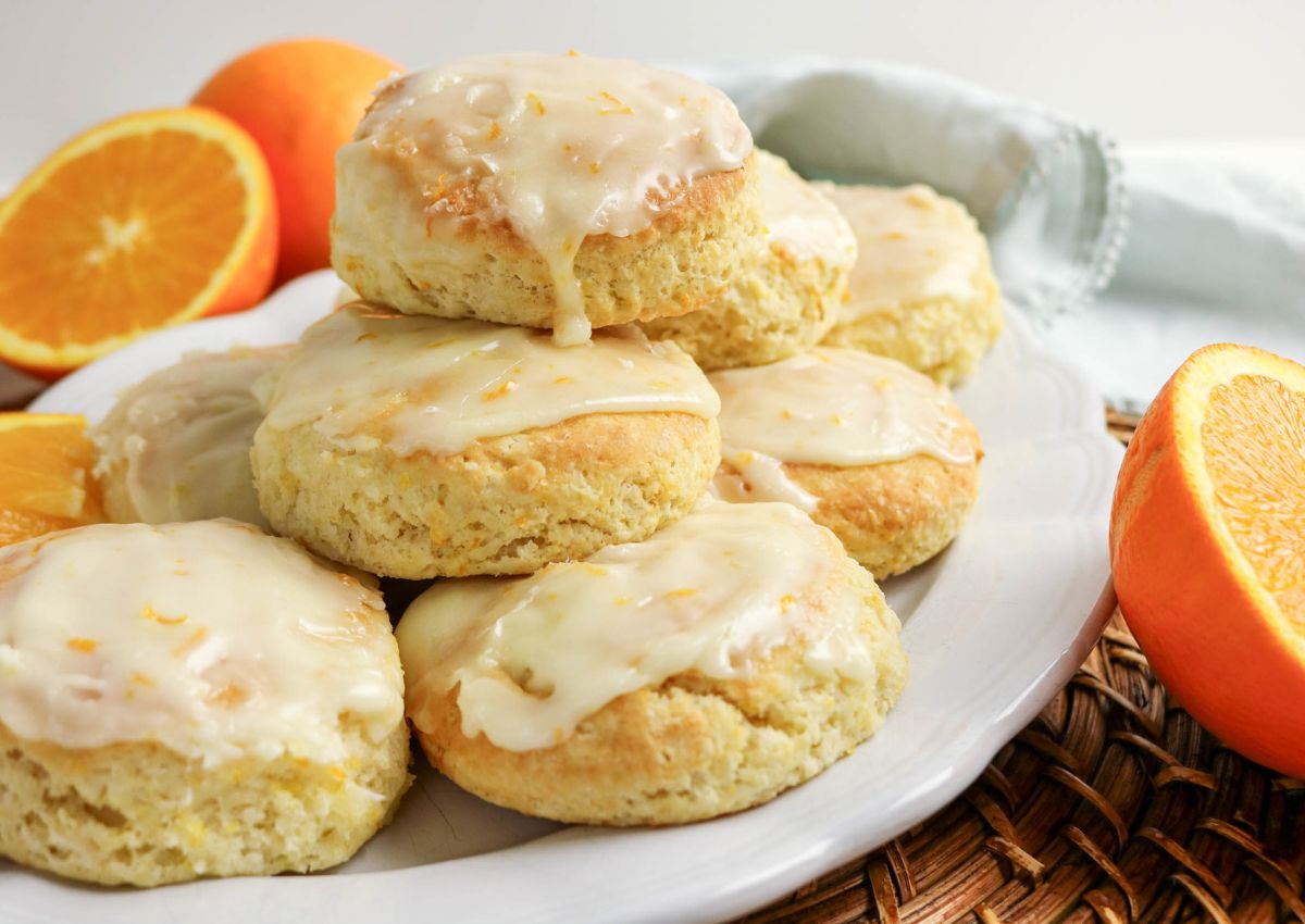 Plate of glazed scones stacked on a white dish, surrounded by halved and whole oranges, on a wicker placemat.