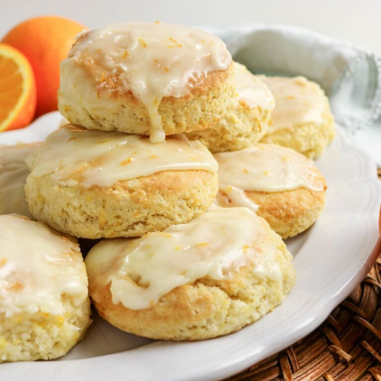 A plate of glazed biscuits with orange zest on top, arranged in a stack. Two orange halves and a cloth napkin are visible in the background.