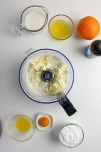 A food processor with flour and butter, surrounded by bowls of milk, melted butter, orange, vanilla, orange zest, orange juice, and powdered sugar on a white surface.