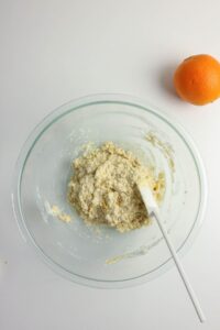 A glass bowl with dough and a white spatula sits on a white surface next to a whole orange.
