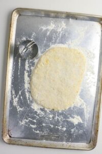 A sheet pan with a floured biscuit dough slab and a metal round biscuit cutter next to it. Flour is scattered on the pan’s surface.