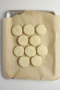 Ten raw biscuit dough rounds are arranged in a rectangle on a parchment-lined baking sheet, ready for baking.
