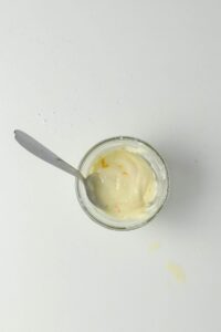 A small glass jar of creamy yogurt with orange zest, partially eaten, and a metal spoon inside, sits on a white surface with a few drops spilled nearby.