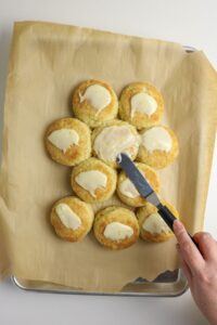 A hand spreads icing on a cluster of round biscuits arranged on parchment paper on a baking tray.