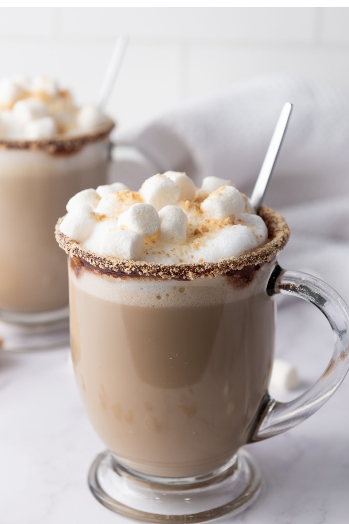 Clear glass mugs filled with a creamy beverage, topped with whipped cream, mini marshmallows, and a graham cracker rim, with spoons inserted and another similar mug in the background.