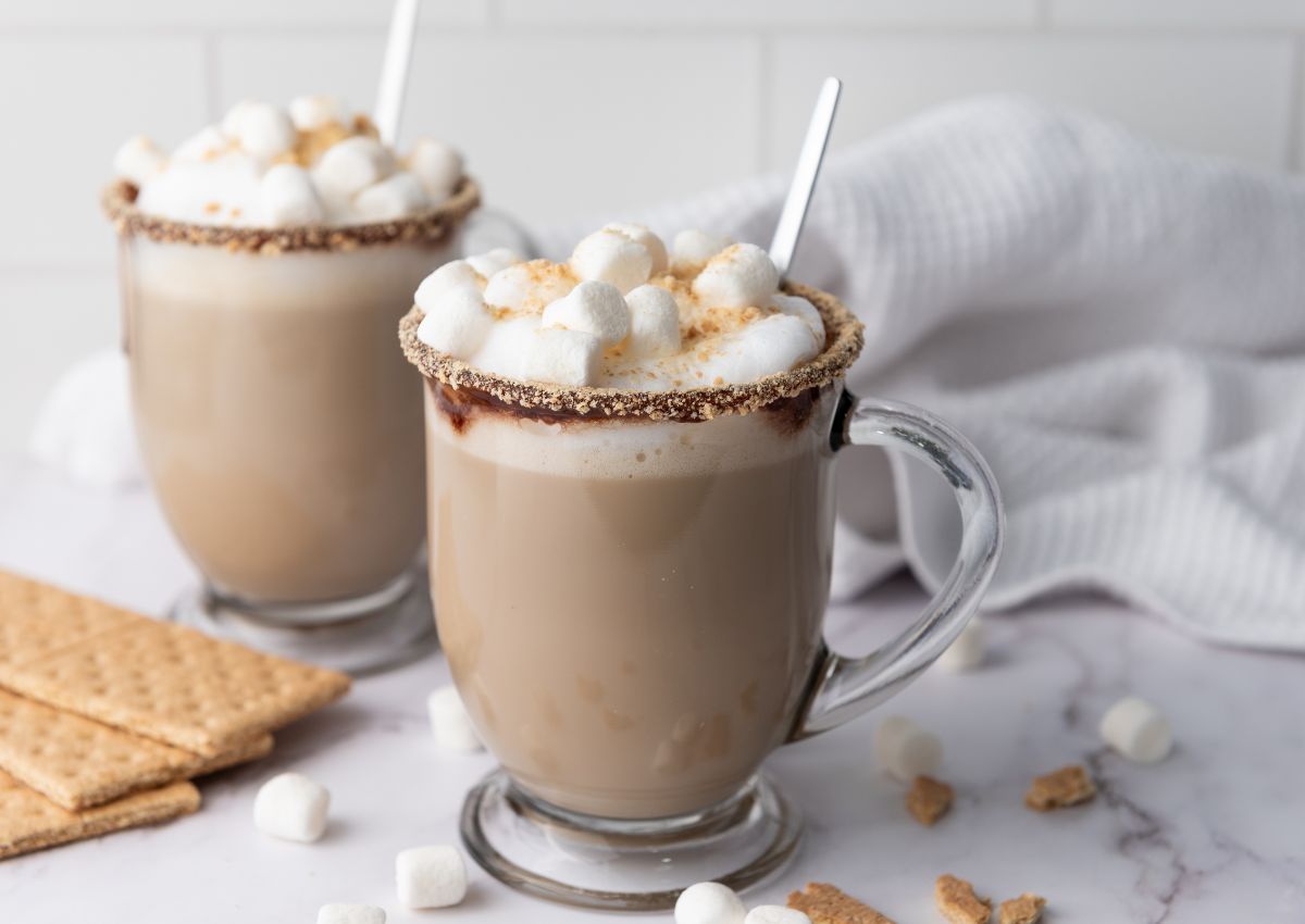 Two glass mugs of hot chocolate topped with mini marshmallows and graham cracker crumbs, with graham crackers and marshmallows scattered on a marble surface.
