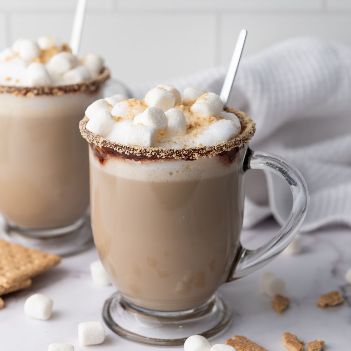 Two glass mugs of hot chocolate topped with mini marshmallows and graham cracker crumbs, each with a spoon, sit on a white surface with scattered graham crackers and marshmallows.