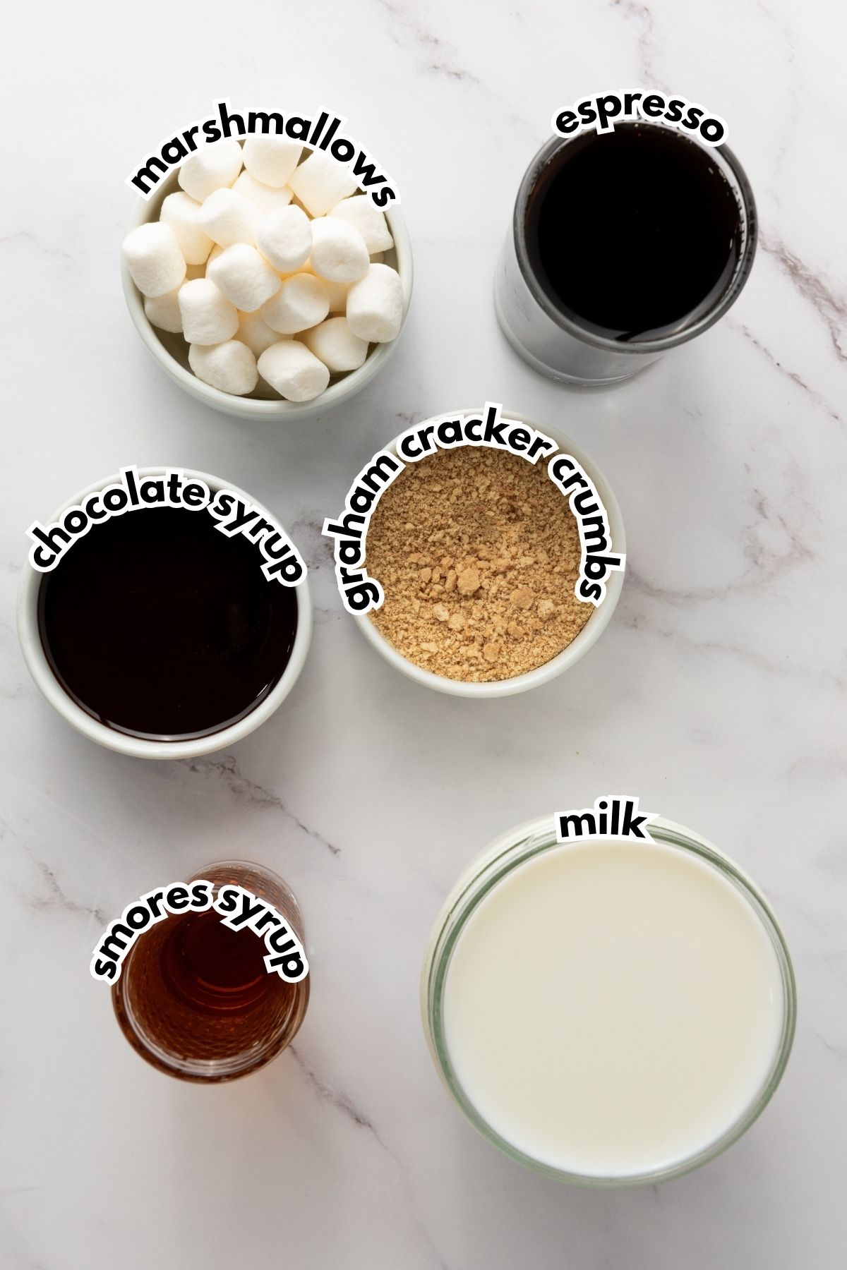 Bowls and cups containing mini marshmallows, espresso, chocolate syrup, graham cracker crumbs, smores syrup, and milk on a white surface with labeled text on each ingredient.