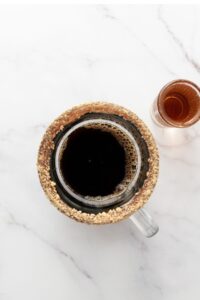 A mug of black coffee with a crumb-coated rim sits next to a shot glass of brown liquid on a white marble surface.