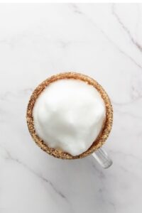 A glass mug filled with a foamy beverage, topped with thick milk froth and a rim coated in crushed crumbs, on a white marble surface.