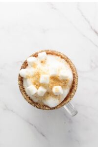 A glass mug filled with a frothy drink topped with mini marshmallows and sprinkled crumbs, on a white marble surface.
