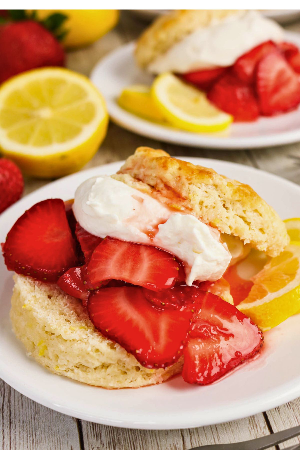 A biscuit topped with sliced strawberries and whipped cream is served on a white plate with lemon slices.