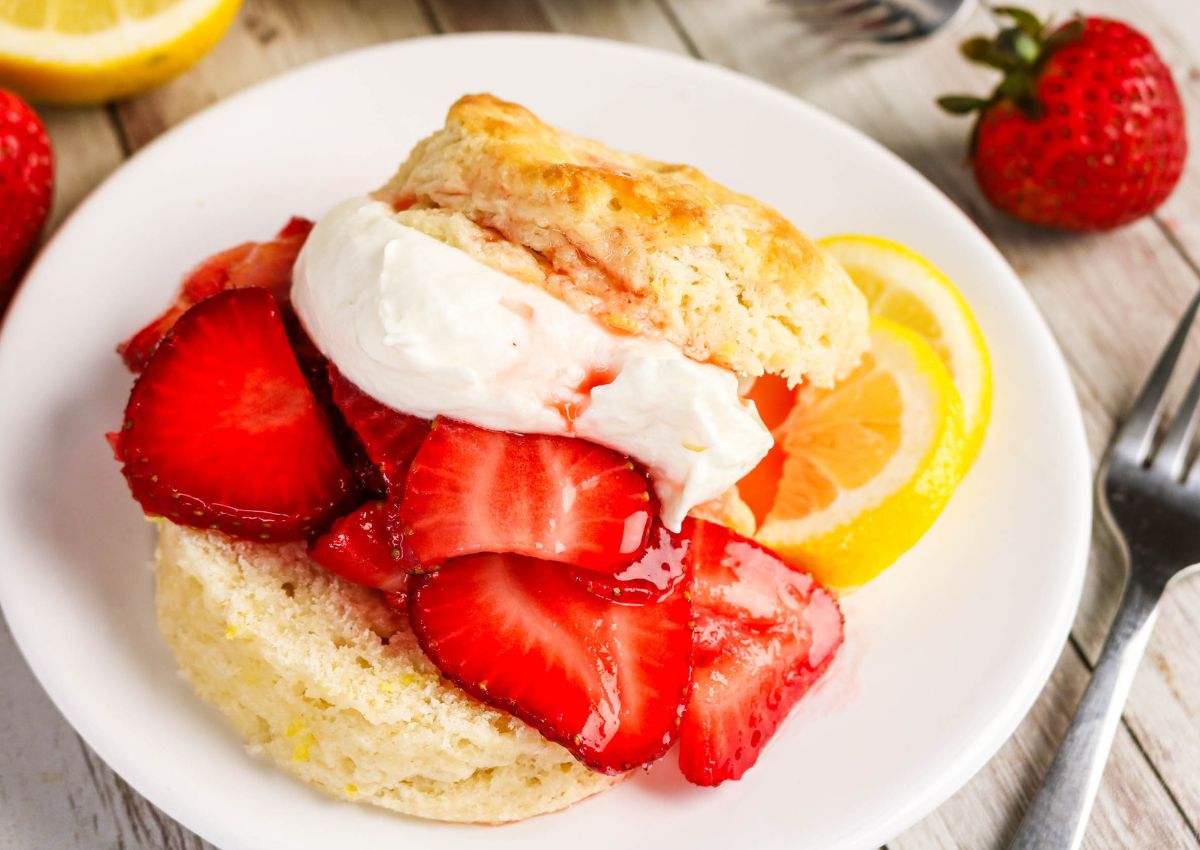 A biscuit split in half, topped with sliced strawberries and whipped cream, is served on a white plate with a lemon slice and whole strawberries nearby.