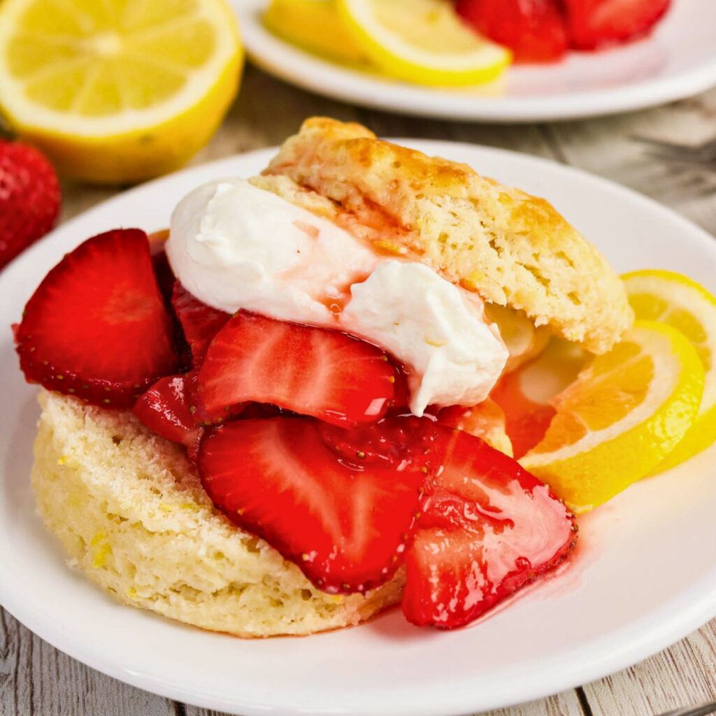 A biscuit split open and topped with sliced strawberries, whipped cream, and lemon slices on a white plate.