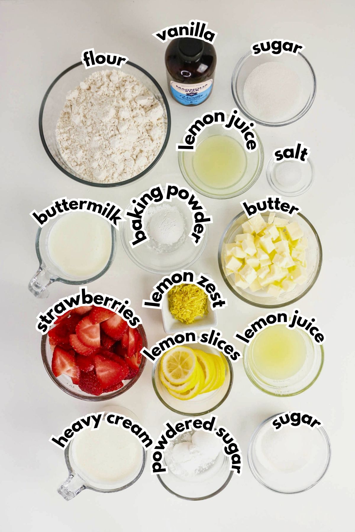 Overhead view of labeled baking ingredients in bowls, including flour, sugar, butter, buttermilk, strawberries, heavy cream, vanilla, lemon juice, lemon zest, lemon slices, baking powder, and salt.