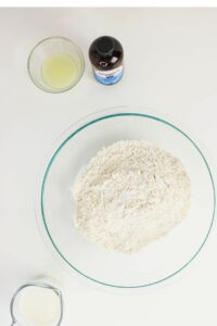 A glass bowl filled with flour, a small bowl of lemon juice, a bottle of vanilla extract, and a measuring cup of milk on a white surface.