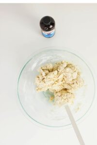 A bowl of dough with a spatula next to a bottle of vanilla extract on a white surface.
