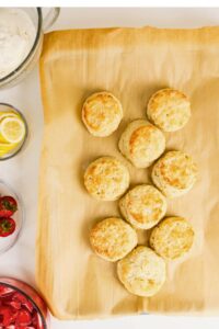 Nine golden biscuits on parchment paper, surrounded by bowls of lemon slices, strawberries, and whipped cream on a white surface.