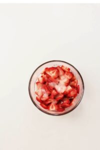 A glass bowl filled with sliced strawberries topped with a sprinkling of sugar, placed on a plain white surface.