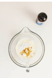 A glass mixing bowl with cream, powdered sugar, and vanilla extract on a white surface; a bottle of vanilla extract is nearby.