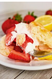 A fork holds a bite of strawberry shortcake with whipped cream, fresh strawberries, and a biscuit on a white plate. Lemons and strawberries are in the background.