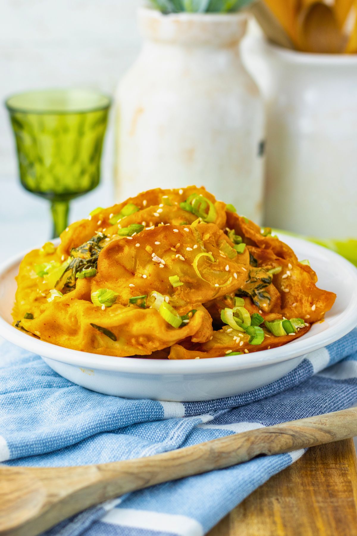 A plate of dumplings topped with chopped green onions and sesame seeds sits on a blue-striped cloth, with a wooden spoon and glassware in the background.