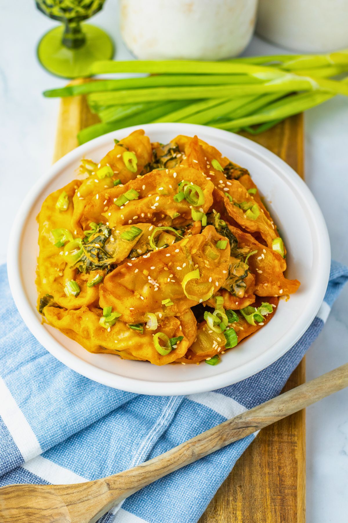 A white bowl of dumplings topped with sliced green onions and sesame seeds sits on a wooden board, with fresh green onions and a blue cloth nearby.