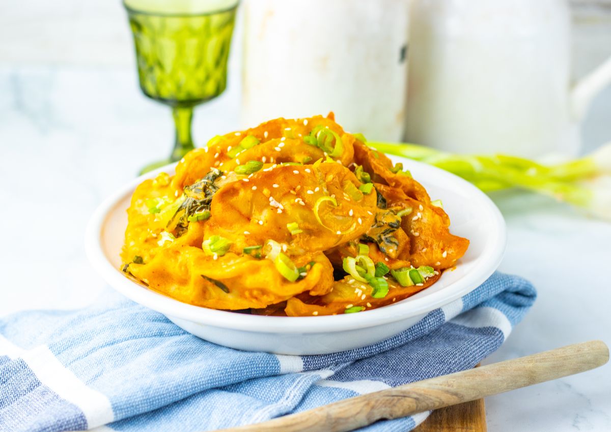 A white bowl filled with kimchi garnished with chopped green onions and sesame seeds sits on a blue cloth napkin with a wooden spoon nearby.