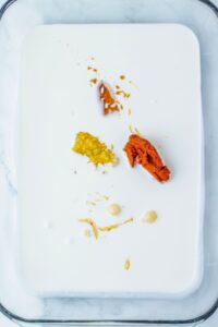 A glass dish filled with white liquid, topped with small portions of red paste, yellow paste, brown liquid, and beige dollops.