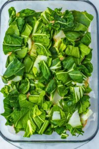 Chopped leafy greens evenly spread over a layer of ingredients in a clear glass baking dish, ready for cooking.